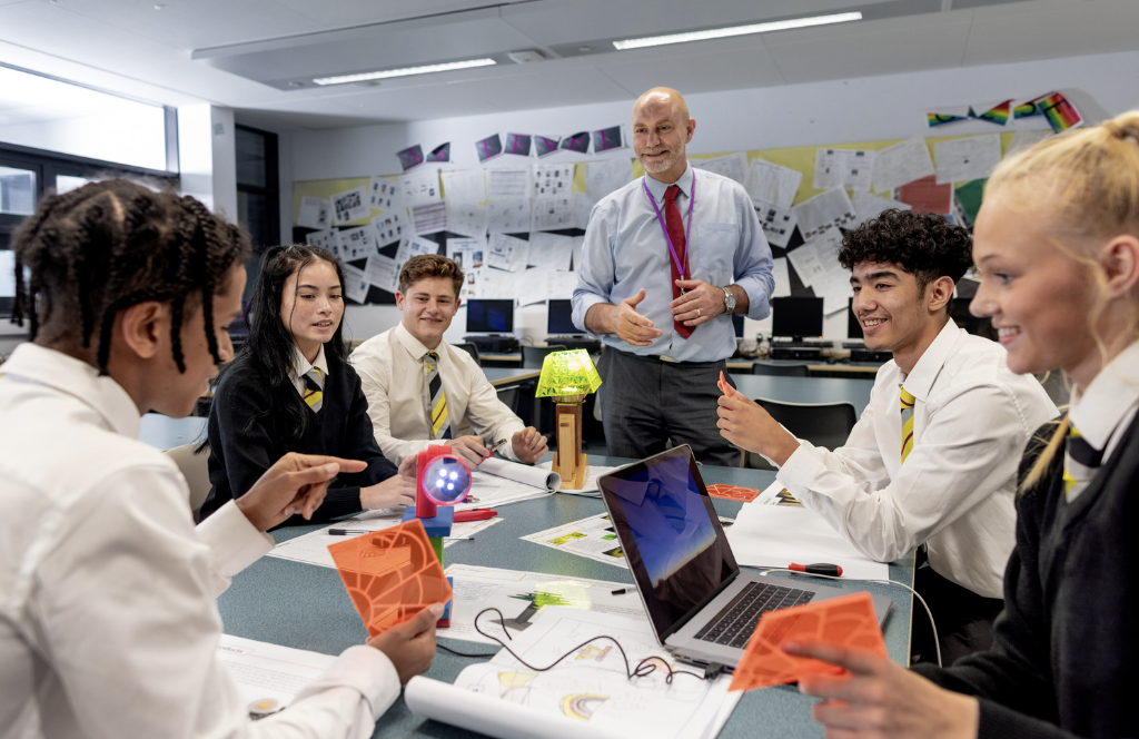 Image of students in a classrrom around a desk with a teacher at the front of the desk.